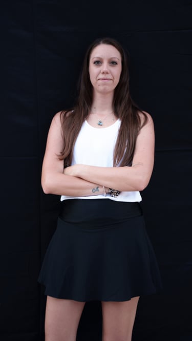 A young woman posing with arms crossed wearing a white tank top and black athletic skirt.