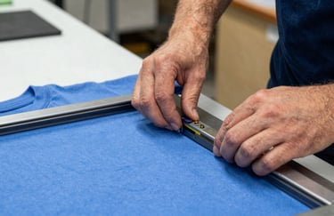 Close-up of a technician's hands carefully aligning a screen-printing frame over a blue cotton t-shirt in a workshop.
