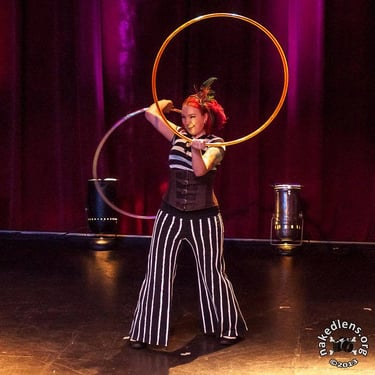 A smiling woman dancing with 2 shiny hula hoops in a direct spotlight on stage