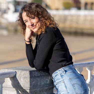 Smiling woman with curly hair wearing a black sweater and blue jeans leaning on a stone railing outdoors.