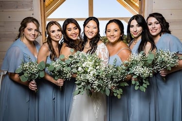 A smiling bride in a lace wedding dress stands next to her bridesmaid in a blue gown holding bouquets.