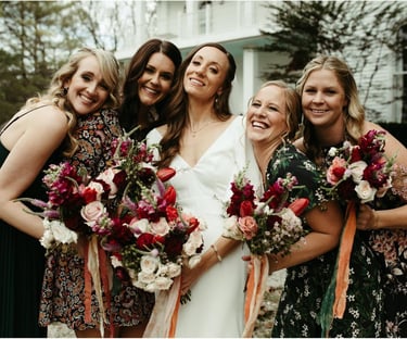 Smiling bride and bridesmaids holding red and pink floral bouquets at a wedding celebration.
