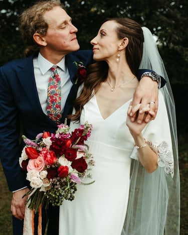 A bride in a white lace gown and groom in a navy suit holding a red rose wedding bouquet.