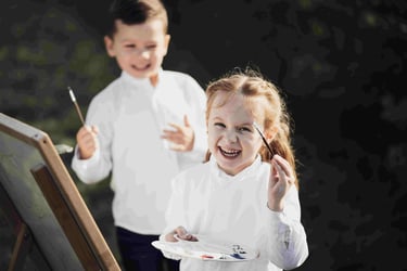 Two kids, wearing white shirts, happily paint outdoors.