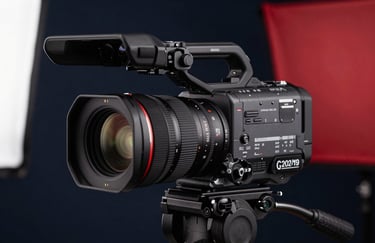 Close-up of a high-end digital video camera on a tripod, focus on the lens. The background is a soft blur of a studio with dark navy and deep red light panels.