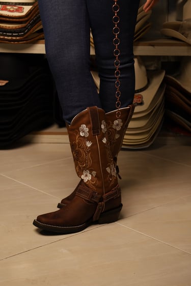 Brown leather women's cowboy boots with white floral embroidery and spur straps worn with blue jeans.