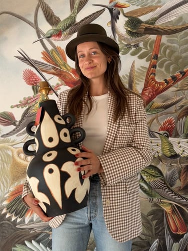 Woman in a blazer holding a large black patterned ceramic vase against hummingbird wallpaper.