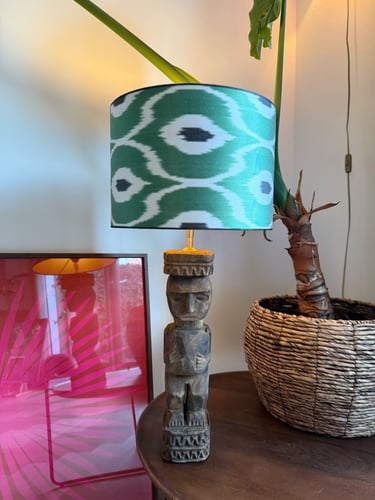 Hand-carved wooden tiki statue lamp with a green ikat pattern lampshade on a wooden table.