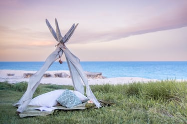 A cozy teepee tent with pillows on a grassy cliff overlooking a beach sunset.