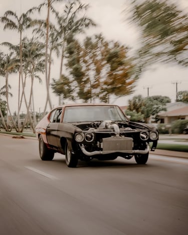 Custom black and red muscle car with exposed turbo engine driving fast past tropical palm trees.