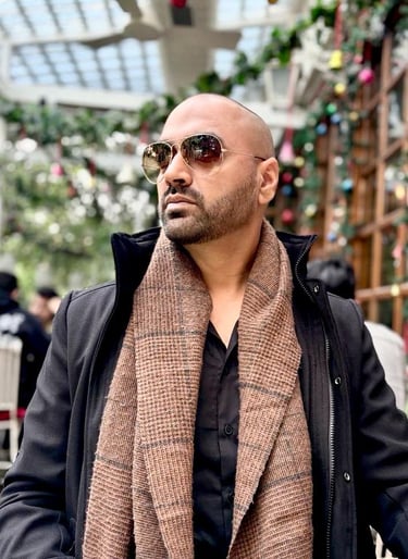 Man with a beard wearing aviator sunglasses, a black jacket, and a brown scarf outdoors.