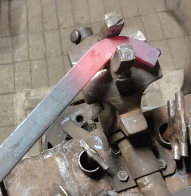 Kalvis lenkia iki raudonumo įkaitintą metalinį strypą, formuodamas saulutės detalę.