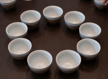 An overhead shot of a coffee cupping session with eight ceramic bowls arranged in a circle on a dark espresso-colored table.