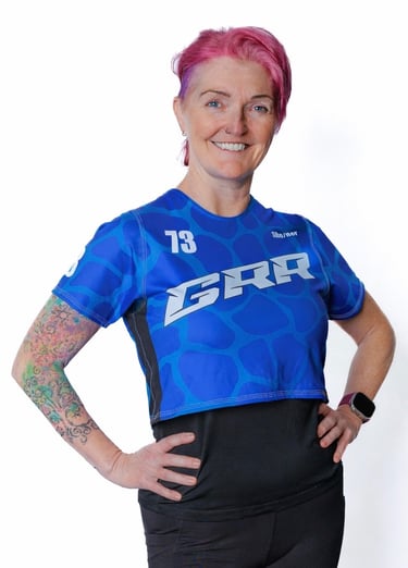 Smiling roller derby athlete with pink hair wearing a blue GRR team jersey and colorful arm tattoo.