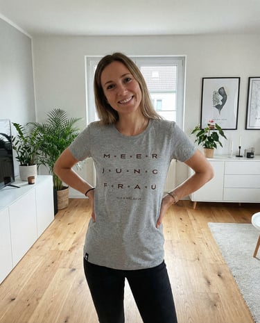Smiling woman wearing a grey Meerjungfrau graphic t-shirt in a modern living room with plants.