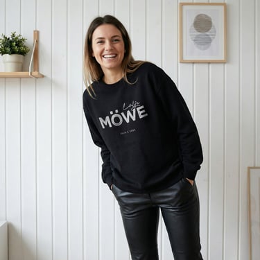 Smiling woman wearing a black Lüttje Möwe sweatshirt and leather pants in a minimalist room.