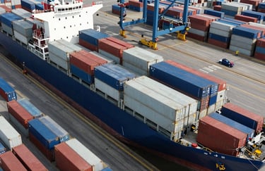 An overhead drone shot of a busy shipping port in the US, showing cargo ships and organized shipping containers in shades of deep blue and light gray.