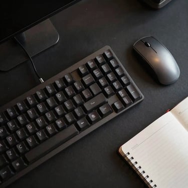 A top-down view of a designer's desk with a dark mechanical keyboard, a silver mouse, and a notebook. Minimalist and dark. North American / International.