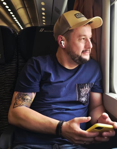 A man with a tattoo and wireless earbuds looking out the window of a Trenitalia train.