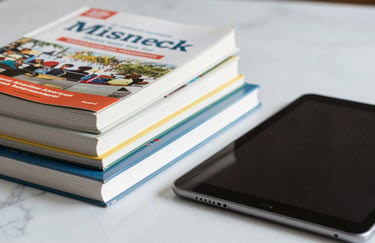 A close-up of premium educational textbooks stacked neatly next to a sleek digital tablet on a white marble surface. North American / International.