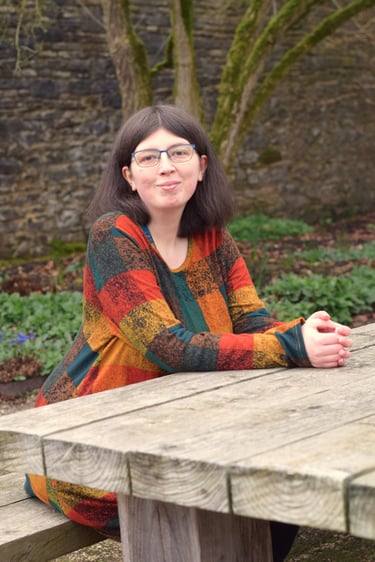 Bex sitting at a wooden picnic table in a garden, wearing a colourful patchwork top and glasses