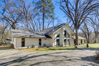 Modern white ranch house with large windows and vaulted ceilings surrounded by tall oak trees.