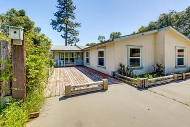 A light yellow ranch-style house featuring a rustic wooden deck and birdhouse in a sunny yard.