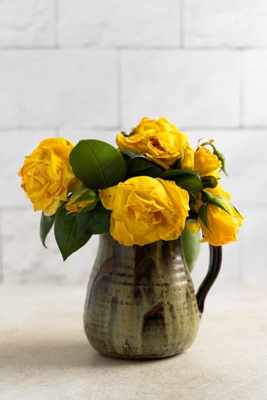 Bouquet of vibrant yellow wilted roses in a rustic ceramic pitcher vase against a white brick wall.