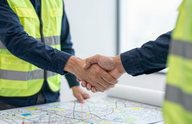 A close-up shot of two professionals in safety gear shaking hands over a set of detailed site maps. The lighting is bright and clear, emphasizing trust and professional partnership, with brand colors #1A202C and #F7FAFC.