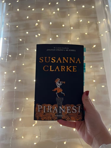 Mână ținând cartea 'Piranesi' de Susanna Clarke pe un fundal de luminițe de basm.