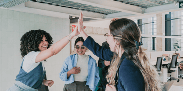 Diverse professional women celebrating success with a high five in a modern office workspace.