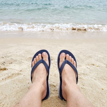 Person wearing navy blue flip flops on a sandy tropical beach with ocean waves.