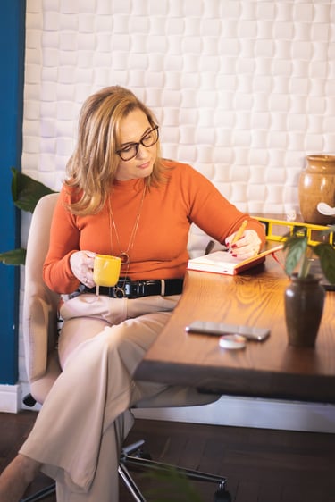 A professional woman in an orange sweater takes notes at a wooden desk with a yellow coffee mug.
