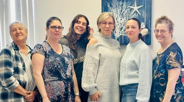 Six smiling professional women posing together in an office with coastal decor.