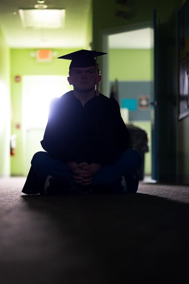 Graduate sitting in a hallway backlit by the sun coming in through the door window