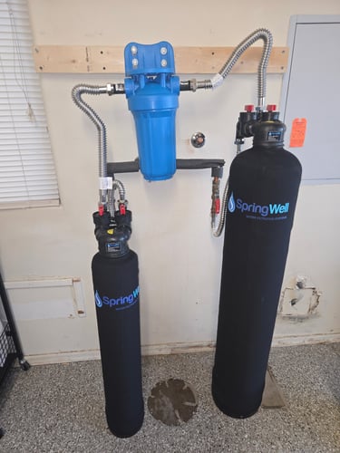 A wall-mounted SpringWell whole house water filtration system with blue sediment filter and two black tanks.