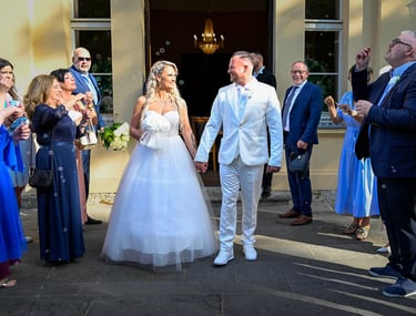 Braut und Bräutigam verlassen das Gebäude nach der Trauung, umgeben von Hochzeitsgästen mit Seifenblasen.