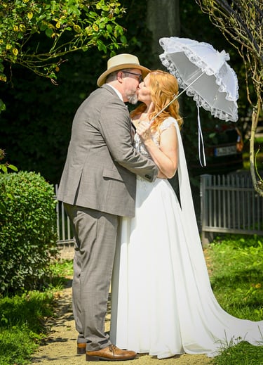 Küssendes Brautpaar im Grünen mit weißem Spitzenschirm nach der Hochzeit.