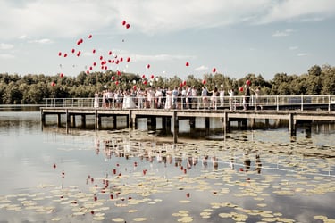 Hochzeitsgesellschaft lässt rote Luftballons auf einem Steg am See steigen – Gruppenmoment einer Hochzeitsreportage.