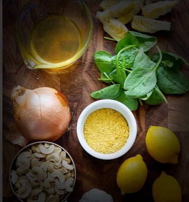 Food prep of onion, spinach leaves, lemons, cashews, artichokes, garlic on chopping block.