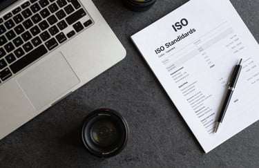A top-down view of a professional desk with a laptop, an ISO standards manual, and a pen. Elegant corporate aesthetic with dark gray and light gray tones, very organized.