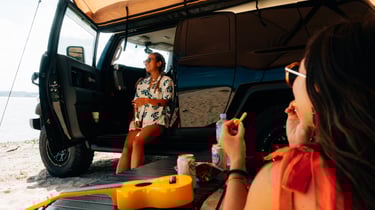 Two women enjoying snacks and drinks beside their Toyota FJ Cruiser parked on the beach in Okinawa. | Evertrail Okinawa