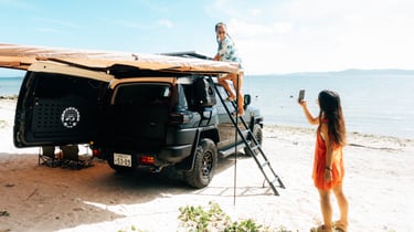 Two friends set up camp at the beach. One climbs the ladder on the black Toyota FJ Cruiser, while the other takes a photo.