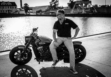 Ein Schwarz-Weiß-Foto eines Mannes, der auf einem Motorrad sitzt und über einen Gewässer blickt. Der Mann trägt eine Sonnenbr