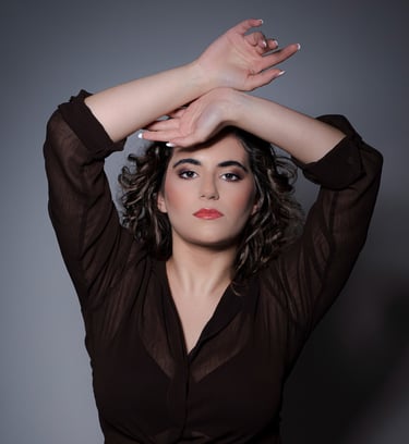 Ein Porträt einer Frau mit dunklen, lockigen Haaren und roten Lippen. Sie trägt eine durchsichtige, dunkelbraune Bluse und ha