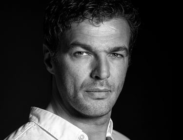 Ein Schwarz-Weiß-Porträt eines Mannes mit dunklem, lockigem Haar und einem leichten Bart. Er trägt ein helles Hemd und blickt