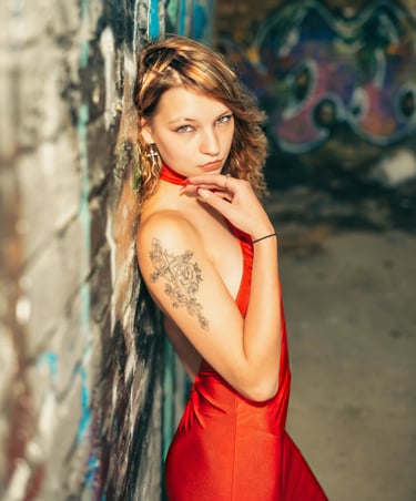 Junge Frau im roten Kleid mit Tattoo am Arm lehnt an einer Graffiti-Wand und blickt direkt in die Kamera – urbanes Portrait v
