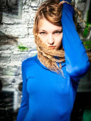 Künstlerisches Portrait einer jungen Frau mit blondem Haar im leuchtend blauen Kleid, intensiver Blick in die Kamera vor urba