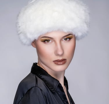  Porträt einer Frau mit weißer Fellmütze, dunkler Bluse und auffälligem Make-up.