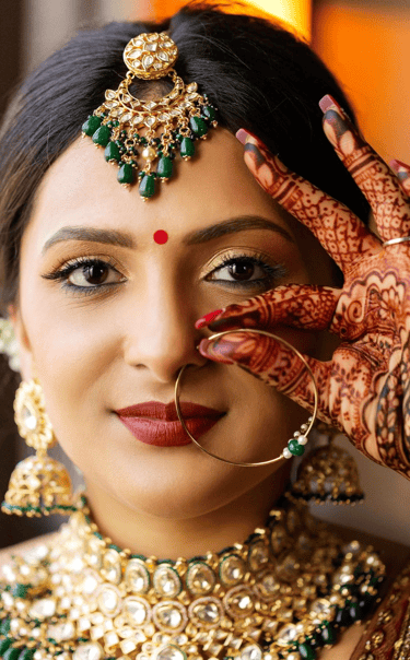Close-up of an Indian bride with soft-glam wedding makeup, red bindi, nose ring, ornate gold-and-emerald jewelry, and henna.